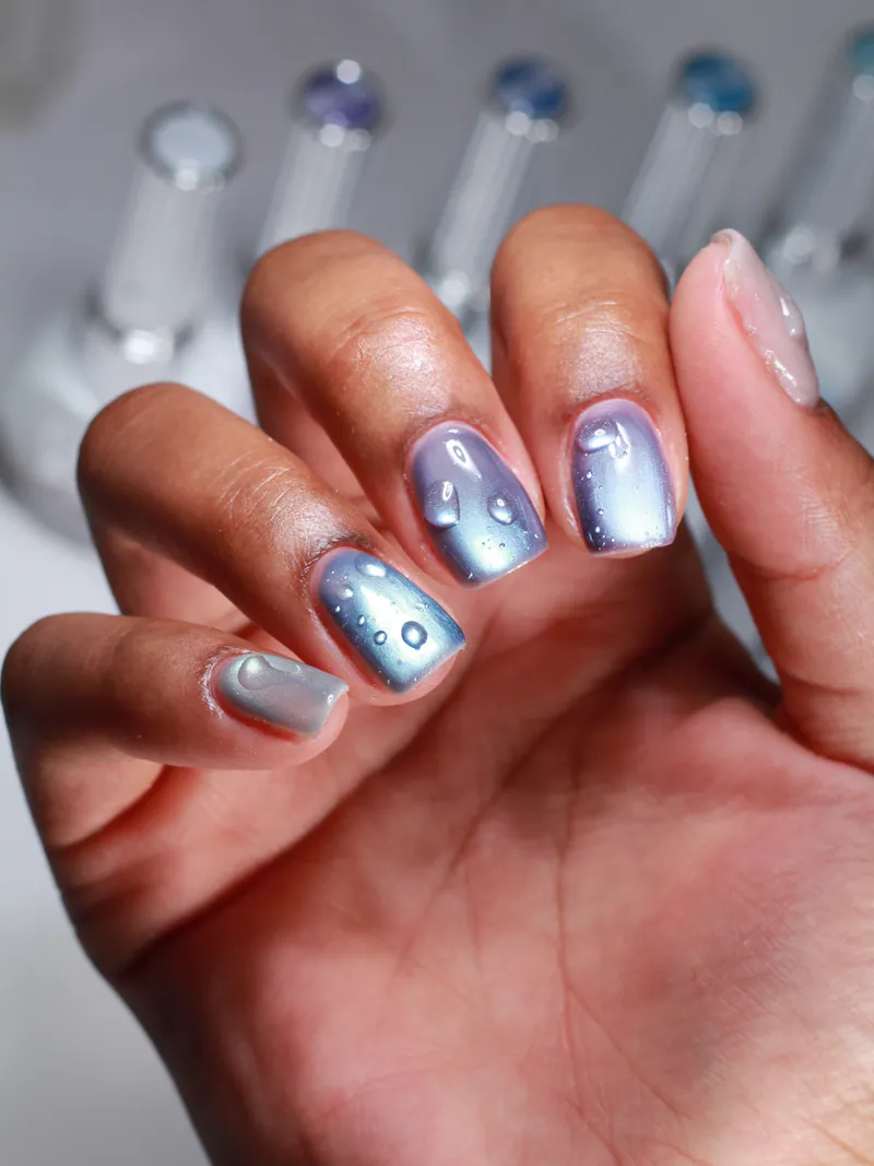 A hand with short square nails in pale silver-blue cat-eye with scattered raised clear droplets, gel bottles blurred behind.