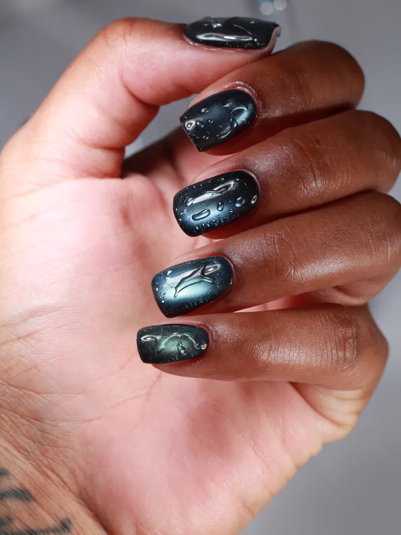 A hand with short square nails in black-teal cat-eye under clear raised droplets.