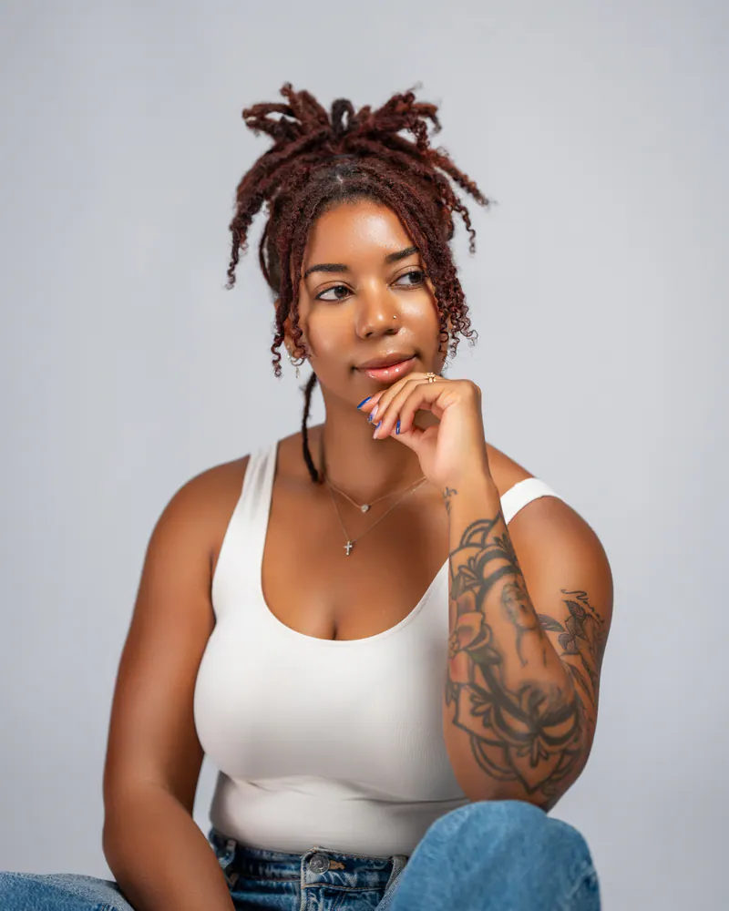 Kazani Dunn in a white tank top, resting her chin on her hand, her tattooed forearm and nails in view.