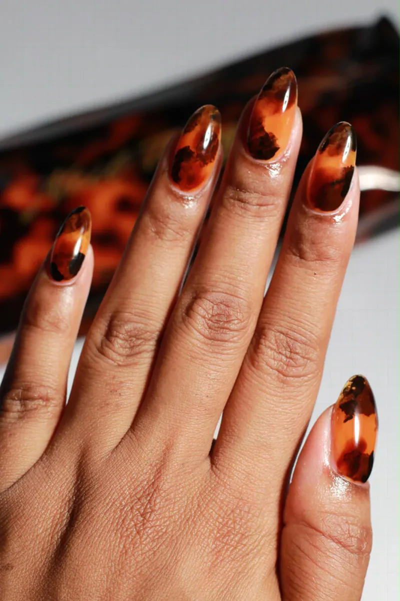 Kazani’s hand with long almond nails in glossy tortoiseshell, warm amber with dark brown patches, a tortoiseshell case blurred behind.